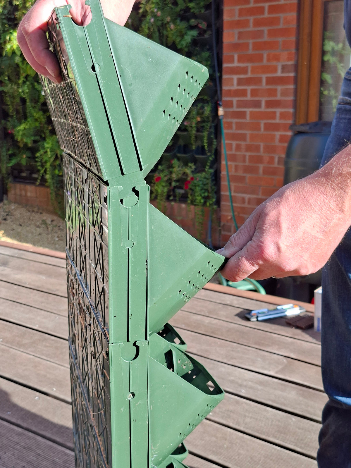 A person's hands assembling a green metal garden trellis or fence panel outdoors on a wooden deck, with a brick wall and plants in the background.